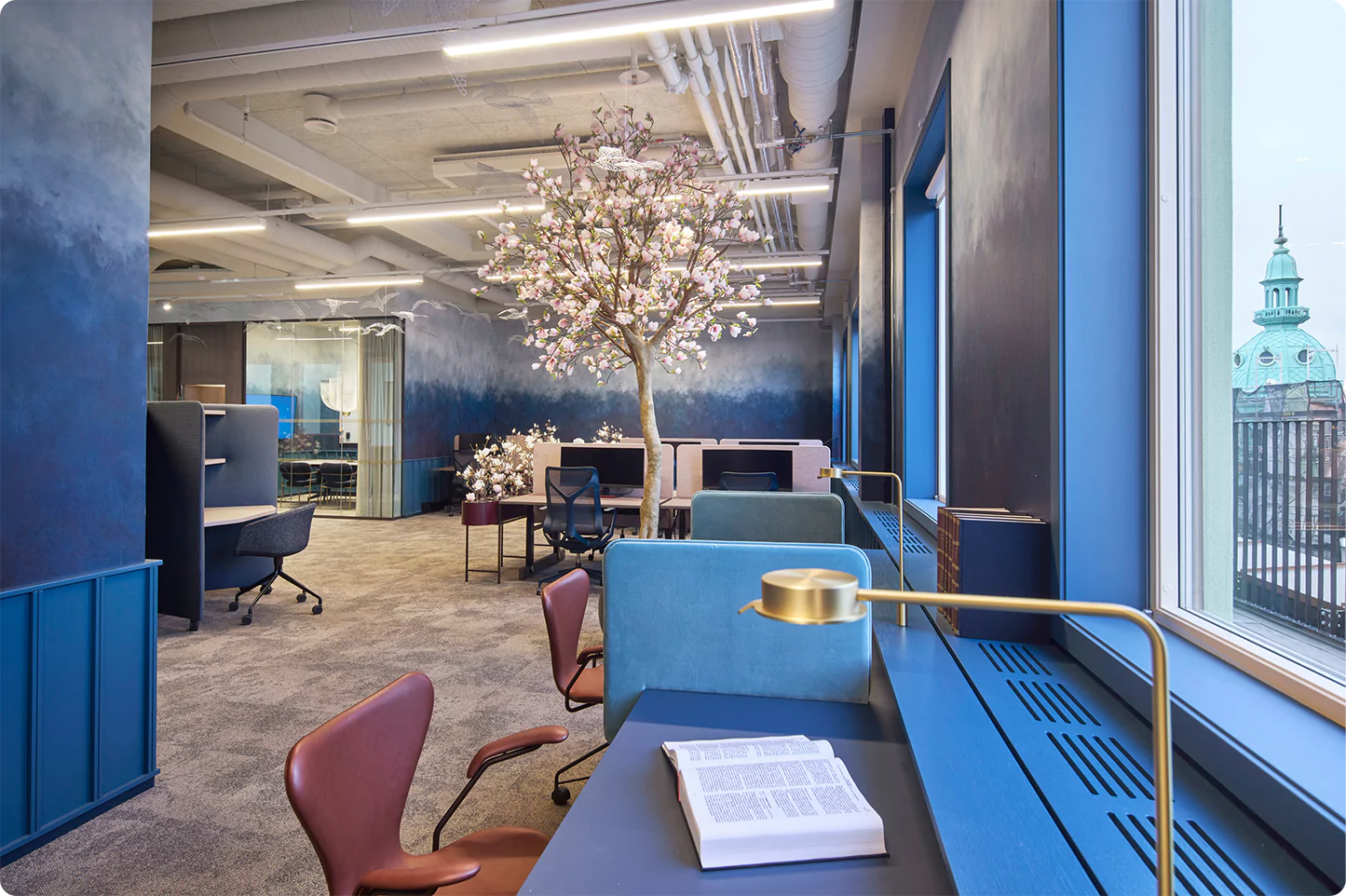 Bilde fra EYs kontor på stortorvet 7 i oslo. bildet går i blå lune farger og midt i rommet står et magnoliatre. det er noen enkeltplasser der man kan sitte med eget arbeid og litt lenger bort i høyre hjørne ser man et møterom.